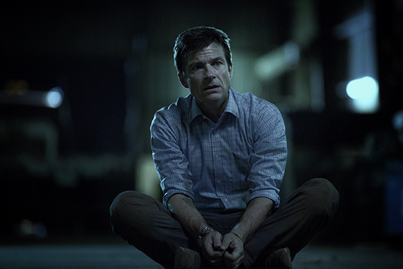 A man in a light blue checkered shirt sits cross-legged on the ground in a dark, dimly lit room.