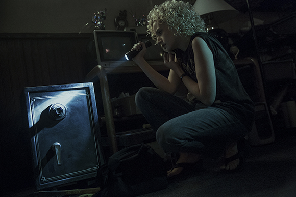 A person with curly hair crouches in a dark room, shining a flashlight on a small metal safe.