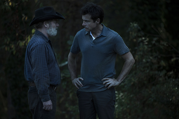 A man in a cowboy hat faces a man in a blue polo shirt in a dark forest.