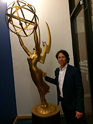 A man in a dark suit stands beside a large, gold Emmy Award statue.
