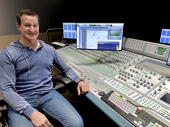 A man in a blue hoodie sits smiling next to a large digital audio mixing desk with multiple monitors.