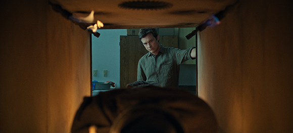 A man looks through a narrow, dark rectangular opening into a brightly lit room.