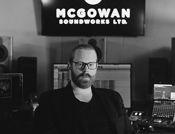 A bearded man wearing glasses and a dark blazer sits in a recording studio in front of computer monitors.