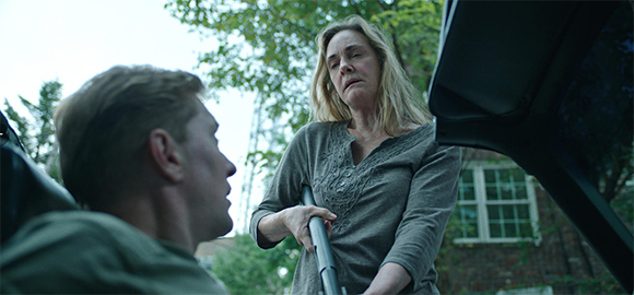 A blonde woman in a grey shirt looks down at a man sitting in a vehicle outdoors.