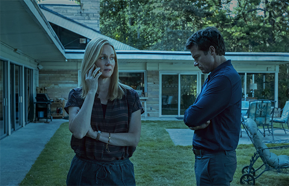 A blonde woman looks concerned while talking on a phone, facing a man with crossed arms in a backyard.