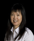 A smiling woman with long dark hair and bangs wearing a white collared shirt against a black background.