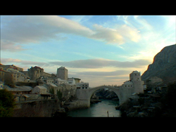 A stone arch bridge crosses a river between rocky cliffs and a hillside town under a cloudy sky.