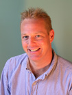 A smiling man with short blond hair wearing a light blue button-down shirt against a teal background.
