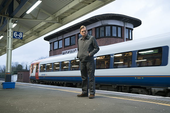 A man in a grey jacket and trousers stands on a station platform next to a white and blue train.