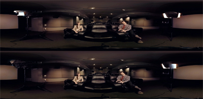A man and a woman sit on a large black sofa in a dimly lit, wide-angle room.