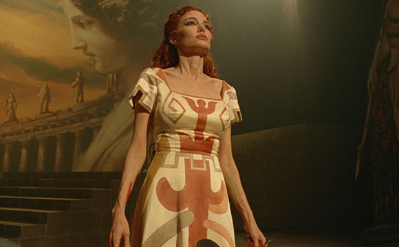 A woman with red hair in a white and terracotta patterned dress stands before a classical mural.