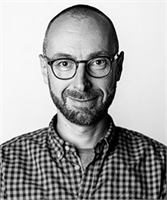 Black and white headshot of a man with short hair, glasses, and a beard wearing a plaid shirt.