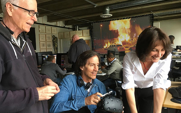 A group of people in a studio look on as a man in a denim jacket holds a VR headset and a smartphone.