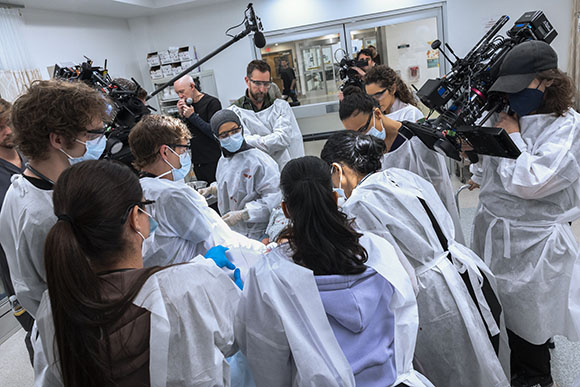 A film crew wearing white protective gowns operates cameras and microphones in a brightly lit room.