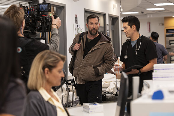 A man in a brown jacket walks through a hospital hallway as a camera crew films him near a medical professional.