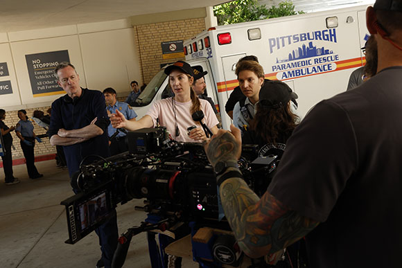 A film crew with a large camera interviews a woman in a pink shirt near a Pittsburgh ambulance.