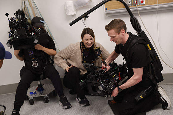 A film crew member adjusts a large professional camera while a woman watches and another person holds a camera rig.