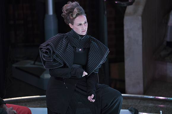 A woman with dark hair in an updo wears a voluminous, structured black garment while sitting on a stage.