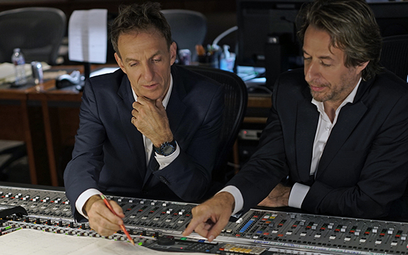 Two men in dark suits lean over a large professional audio mixing console in a studio.
