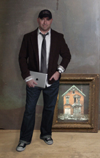 A man in a dark blazer, tie, and jeans stands beside a framed painting of a house.