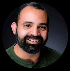 A smiling man with dark hair and a full black beard wearing a green shirt.