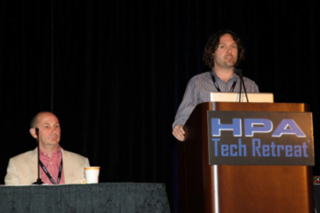 A man stands behind a podium labeled HPA Tech Retreat while another man sits at a table in the foreground.