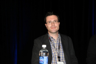 A man with glasses and a plaid shirt under a dark blazer sits before a dark curtain.