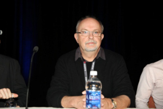 A man with glasses and a goatee wearing a black sweater sits behind a table with a water bottle.