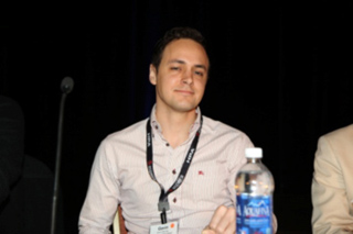 A man in a light-colored button-down shirt sits at a table with a water bottle in front of him.