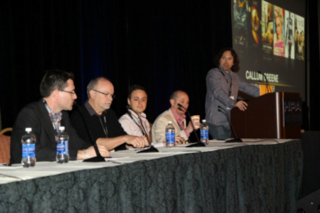 Four panelists sit at a long table while a man stands at a podium next to a screen showing movie posters.