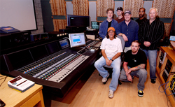Seven men pose together in a music recording studio next to a large mixing console.