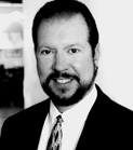 Black and white headshot of a man with dark hair and a beard wearing a suit and tie.
