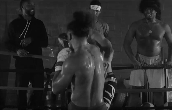 Black and white shot of shirtless boxers and trainers in a boxing ring.