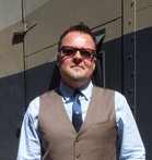 A man wearing sunglasses, a light blue shirt, a blue tie, and a brown vest stands before a dark wall.