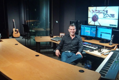 A man smiling while sitting at a desk in a music recording studio with monitors and musical instruments.