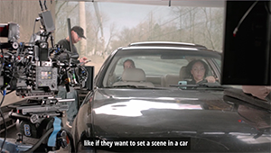 A professional film camera on a rig points toward a dark car with two people visible inside.