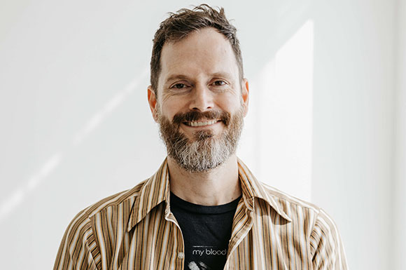 A smiling man with short brown hair and a salt-and-pepper beard wearing a tan striped button-down shirt.