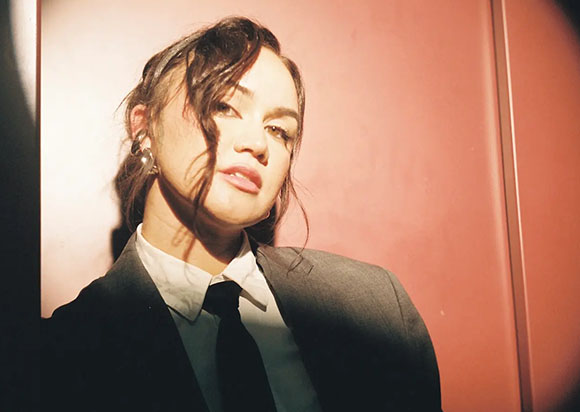 A woman with dark wavy hair wearing a dark suit, white shirt, and black tie against a reddish background.