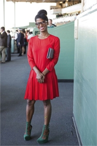 A woman smiling in a long-sleeved red pleated dress and green heels, holding a small clutch.