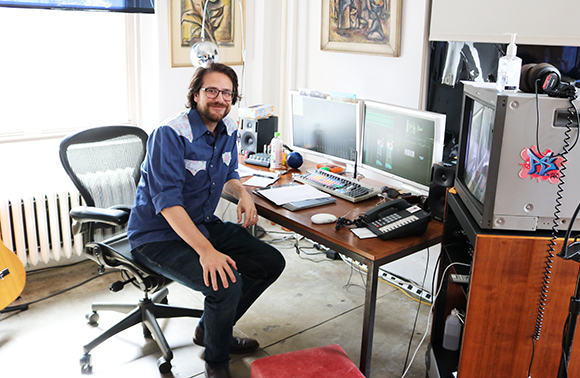 A man with glasses and a blue shirt sits at a desk with computer monitors and audio equipment.