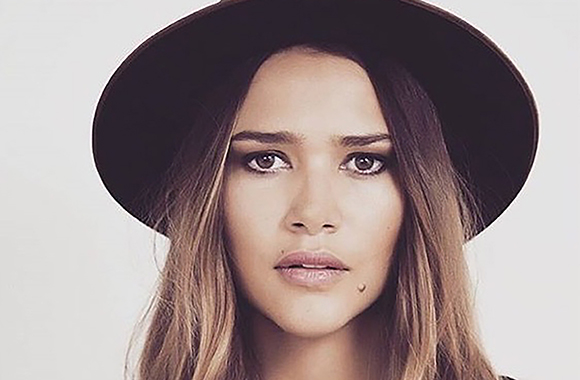 A woman with long brown hair and dark eye makeup wearing a dark wide-brimmed hat against a white background.