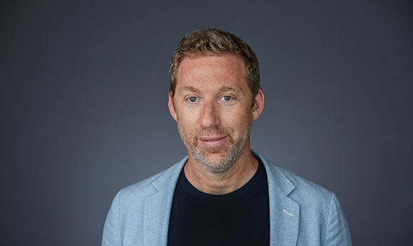 A man with short blond hair and stubble wearing a light blue blazer and black shirt against a dark background.