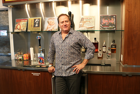 A man in a blue patterned shirt stands behind a stainless steel bar with various bottles and records behind him.