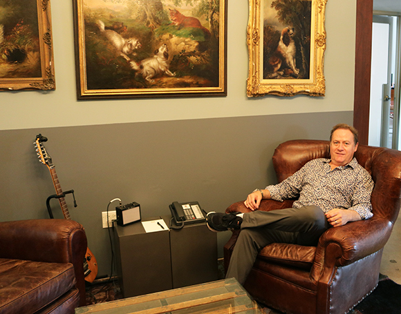 A man in a patterned shirt sits in a brown leather armchair next to a guitar and framed paintings.