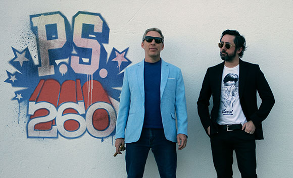 Two men in sunglasses and blazers stand before a blue and red graffiti mural on a white wall.