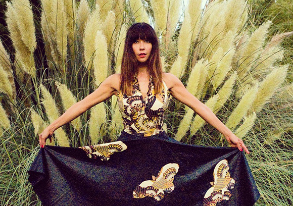 A woman in a patterned halter dress stands with arms outstretched in front of tall, feathery pampas grass.