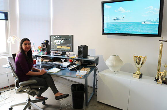 A woman sits at a desk with dual monitors, speakers, and a keyboard in a bright office.