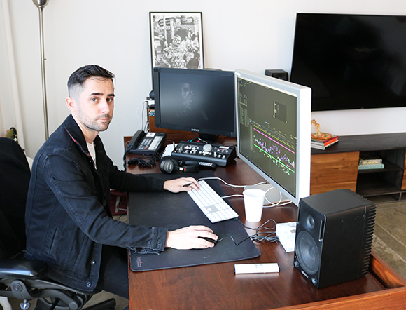A man in a black jacket sits at a desk with two computer monitors, a keyboard, and studio speakers.