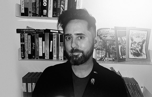 Black and white portrait of a bearded man standing in front of shelves filled with VHS tapes and comic books.