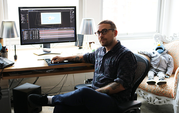 A man with glasses and tattoos sits at a desk using a computer in a bright room.